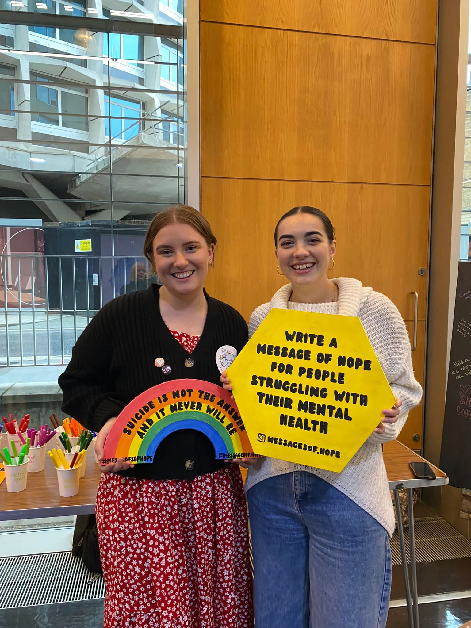 Two of the Hope Bags team at a stall, holding a sign inviting people to write a message of hope for those struggling with their mental health