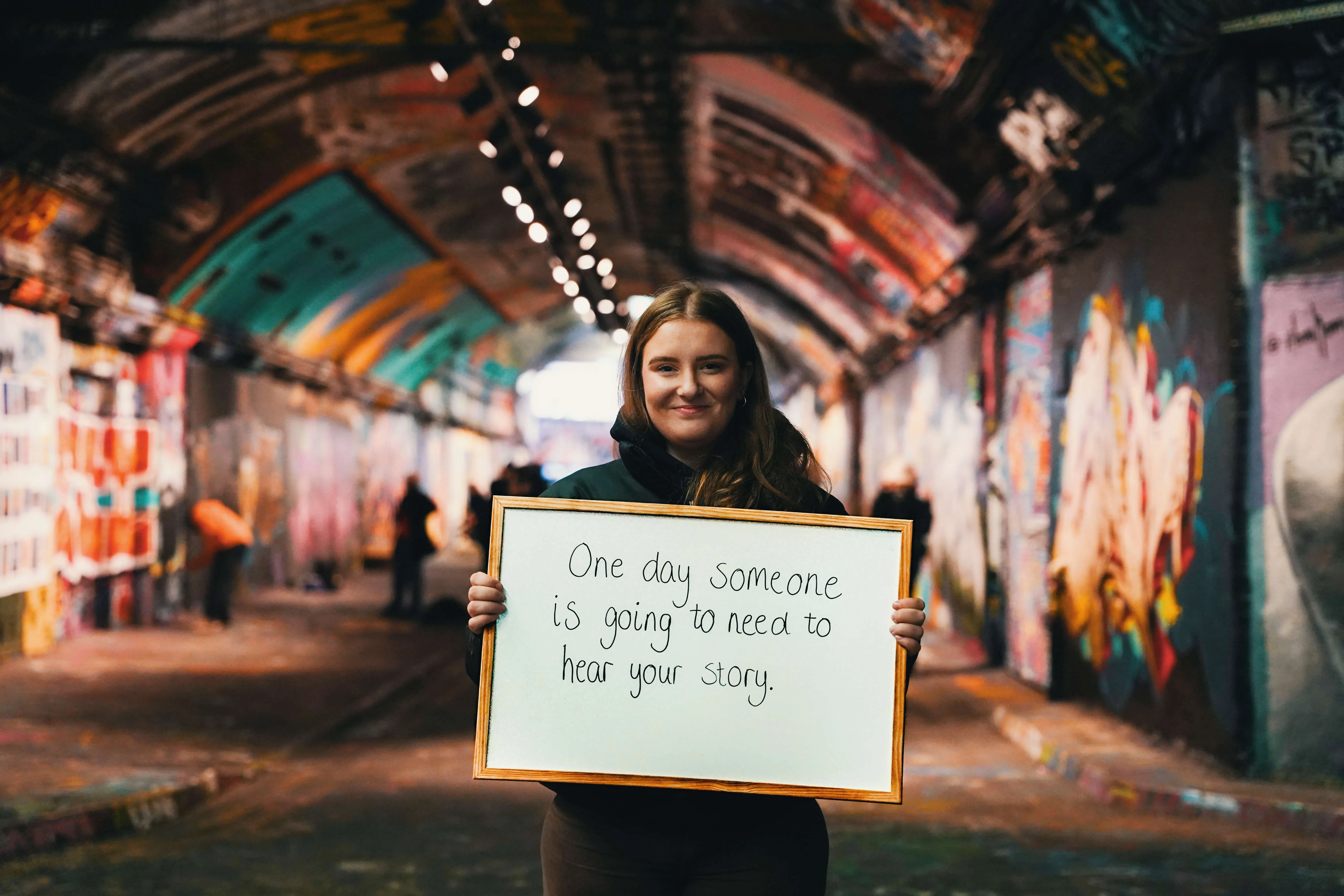 Bethan Evans, founder of Hope Bags, in a graffiti-covered tunnel, holding a handwritten sign that reads 'One day someone is going to need to hear your story.' at Connections 2024